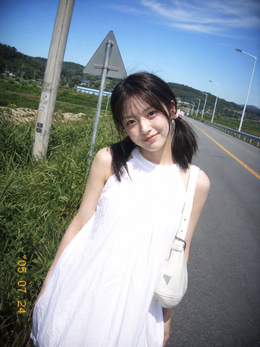 复古 CCD 卡片机夏日旅行抓拍，一位20岁出头的年轻东亚女idol站在乡间公路边。黑棕色低位双马尾，细碎刘海，浅粉色发圈，淡妆，冷白皮，真
